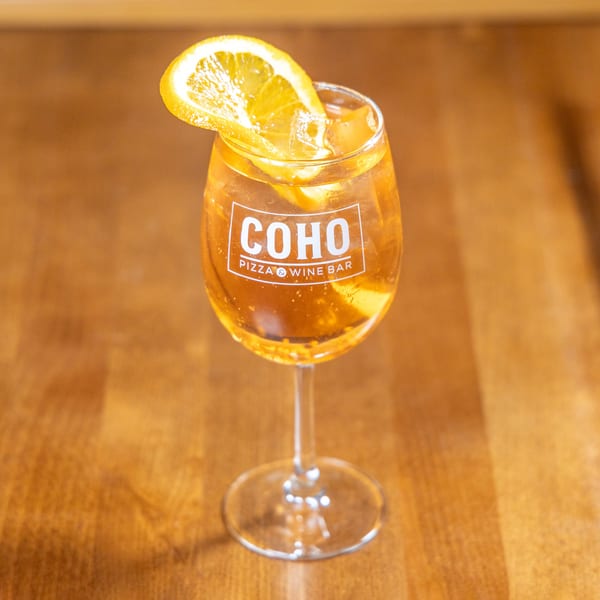 A COHO Pizza & Wine Bar branded glass of orange cocktail with ice and an orange slice.