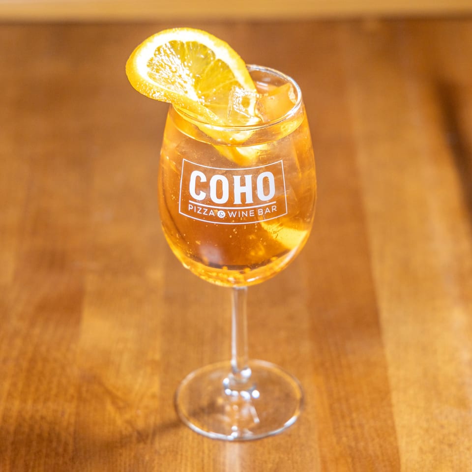 A COHO Pizza & Wine Bar branded glass of orange cocktail with ice and an orange slice.