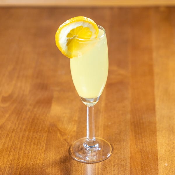 A yellow cocktail in a champagne flute, garnished with a lemon slice, on a wooden table.