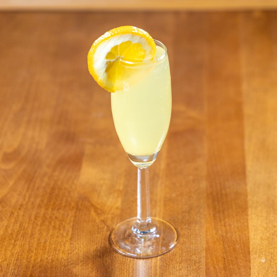 A yellow cocktail in a champagne flute, garnished with a lemon slice, on a wooden table.
