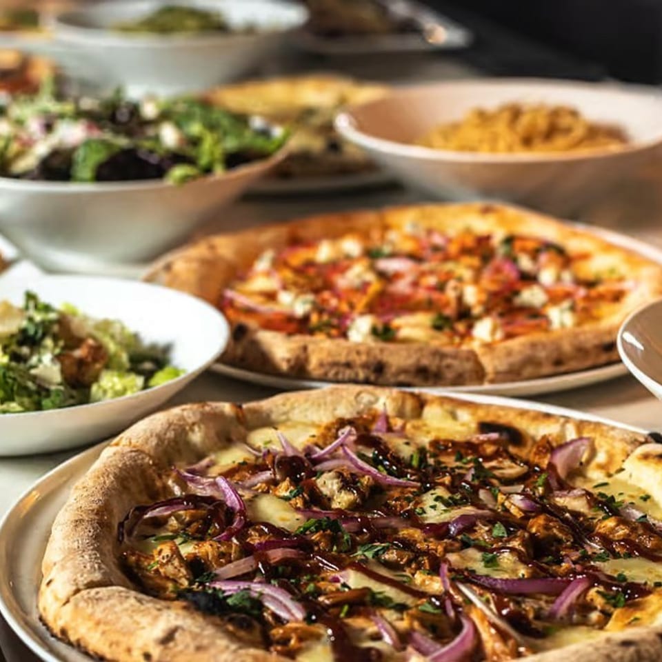 Two COHO pizzas, one with BBQ chicken and red onions, alongside salads and pasta.