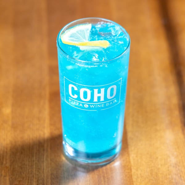 A vibrant blue COHO Pizza & Wine Bar cocktail with ice and a lemon slice on a wooden table.