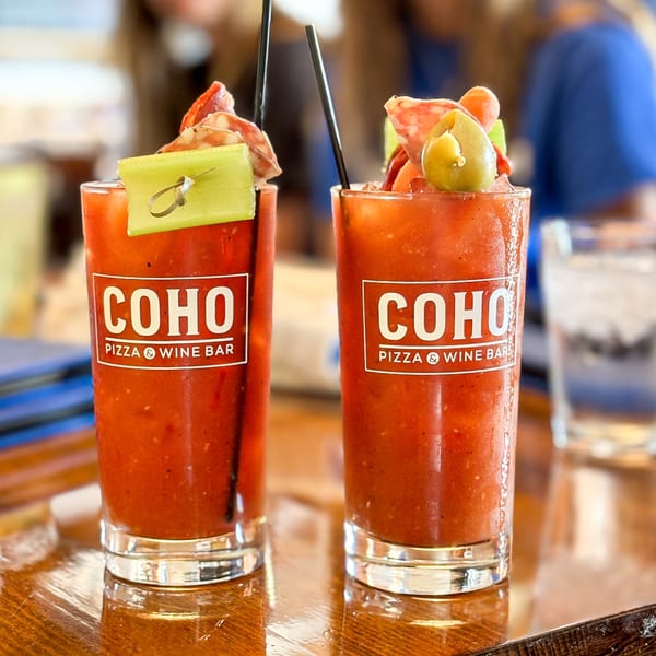 Two Bloody Mary cocktails from COHO Pizza & Wine Bar, garnished with celery, salami, and olives.