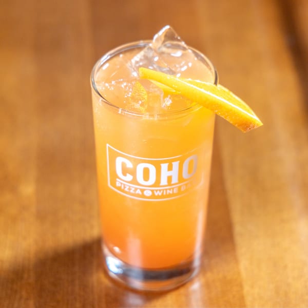 A tall glass of orange cocktail with ice and an orange slice, featuring the COHO logo.
