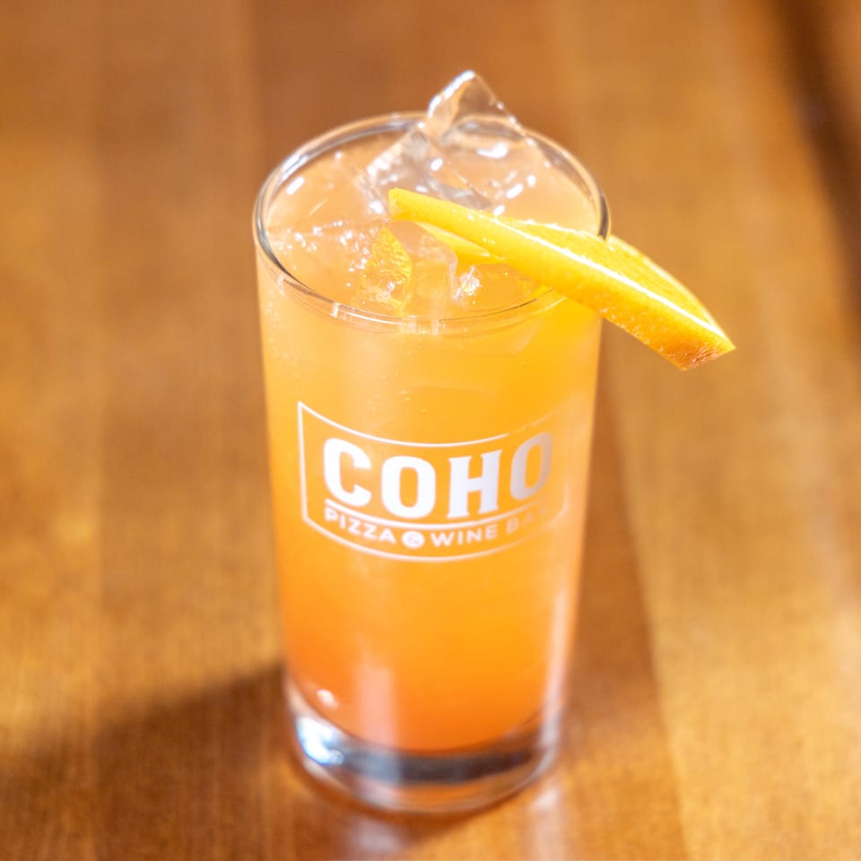 A tall glass of orange cocktail with ice and an orange slice, featuring the COHO logo.