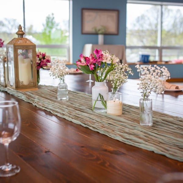 A long wooden table with a sage green runner, decorated with flowers, candles, and a lantern.